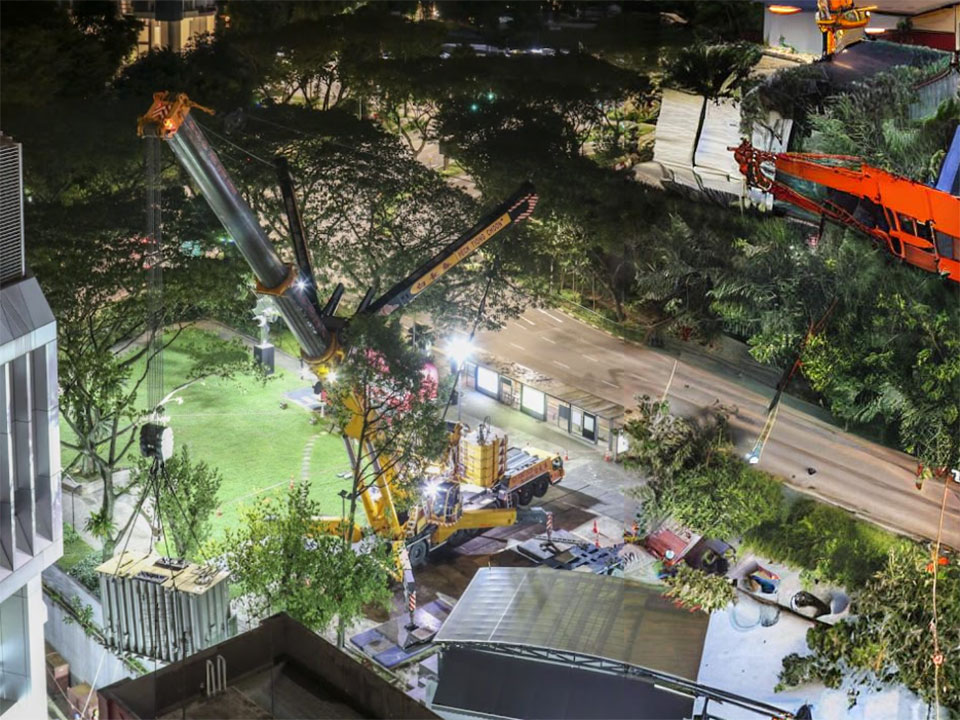 Lifting crane from Peck Tiong Choon carrying container across grass patch during the evening, within a civilian building area.
